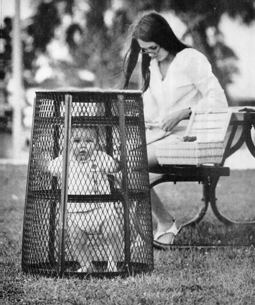 Мама использует мусорное ведро, чтобы содержать своего ребенка, пока она вяжет крючком в парке, 1969 год.